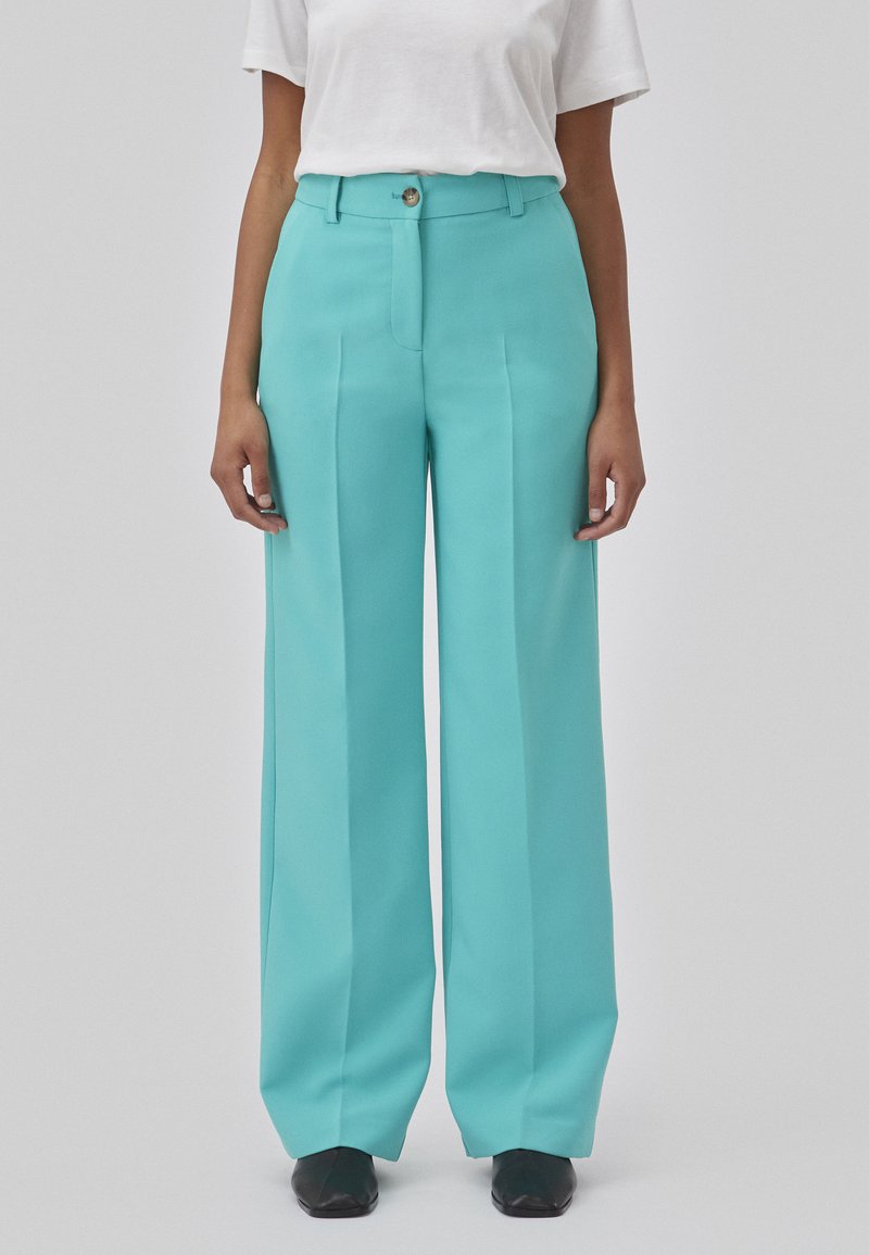 Pantalons larges turquoise en tissu lisse, avec des plis sur le devant et une fermeture à bouton, associés à un t-shirt blanc uni.