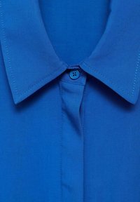 Camicia blu in cotone con una trama liscia, chiusura a bottoni e cuciture a contrasto lungo il colletto.