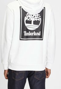 Vit hoodie med stor svart Timberland-logotyp och trägrafik på baksidan, bärs av en person i mörkblå jeans som vänder bort.