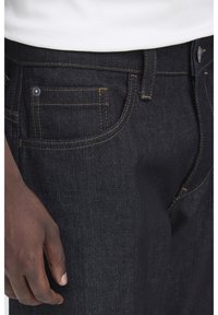 Närbild på mörka jeansfickor och bältesögla med en synlig hand vilande bredvid låret, iförd en vit inskjuten skjorta.