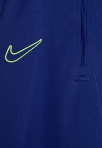 Shorts de sport bleu marine en tissu texturé, avec un logo Nike brodé en vert et une poche zippée sur le côté.