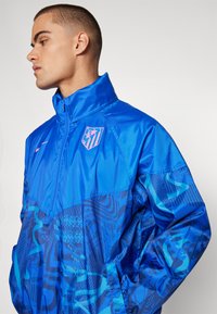 Giacca a vento blu con colletto alto, caratterizzata da un sottile motivo astratto, chiusura con zip e logo del club in rosa sul petto.