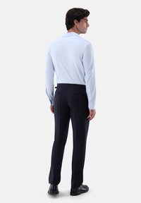 Männlicher Model, der sich abwendet, trägt ein hellblaues Langarmhemd, dunkle Anzughose und schwarze Dressschuhe vor einem schlichten weißen Hintergrund.