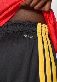 Χέρι που σηκώνει κόκκινη μπλούζα για να δείξει μαύρα σορτς σπορ με κίτρινες ρίγες και λογότυπο "climacool" στην ελαστική ζώνη.