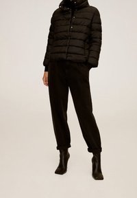 Zwarte pufferjas met een hoge kraag, voorzien van horizontale stiksels en drukknopen, gecombineerd met zwarte manchetten en loszittende donkere broek.