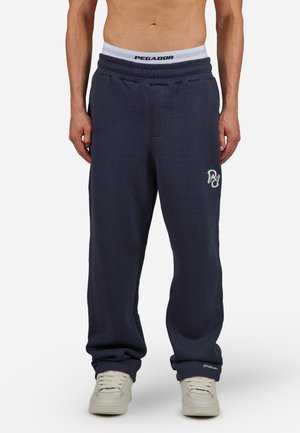Marineblå ribbede joggebukser med elastisk midje og hvit logo-detajle. Taperet passform og myk tekstur, kombinert med hvite sneakers.
