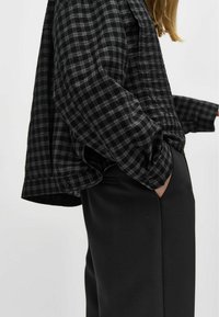 Fekete és szürke kockás flaneling, laza szabású, hosszú ujjú, fekete, zsebbel ellátott, enyhén függőleges motívumú nadrág felett viselve.