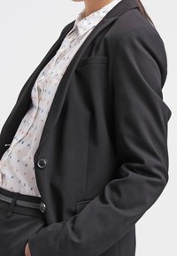Černý upravený blazer se dvěma knoflíky, jednou náprsní kapsou a hladkou texturou. Nosi se přes bílou blůzu s barevnými malými vzory.