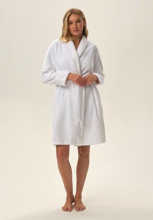 Woman with blonde hair standing barefoot wearing a white plush bathrobe with a textured shawl collar and cuffs.
