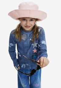 Modrá denimová bunda s farebnými nášivkami hviezd a srdiečok. Svetloružový vedierkový klobúk. Drží čierne slnečné okuliare. Neformálny outfit s voľným strihom.