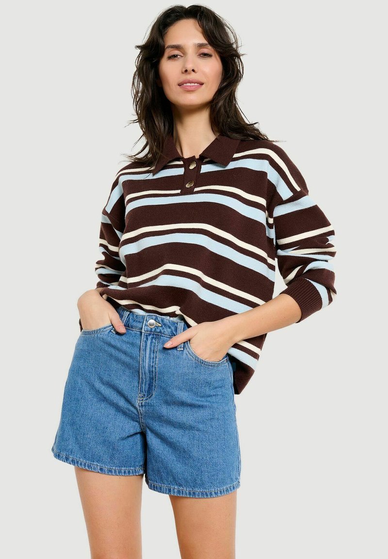 Vrouw die een bruin-wit-lichtblauw gestreept, langarm poloshirt draagt en blauwe spijkershorts, handen in de zakken, tegen een effen achtergrond.