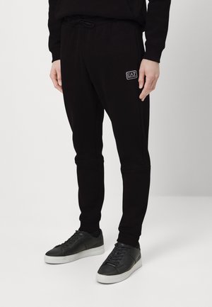 Personne portant un pantalon de jogging noir avec le logo EA7 et des baskets noires à semelles blanches, debout devant un fond blanc uni.