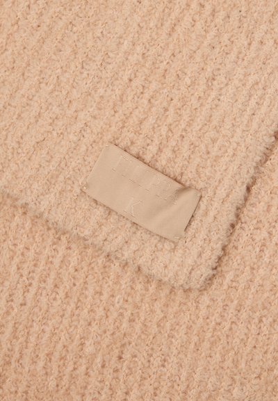 Tejido de punto beige con una superficie suave y texturizada. Presenta una etiqueta con "Filippa K" en relieve, cosida al material.