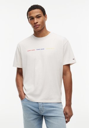 Νεαρός άνδρας που φοράει λευκό T-shirt με στρογγυλή λαιμόκοψη και το κείμενο "Tommy Jeans" με κόκκινα, μπλε και κίτρινα γράμματα, σε συνδυασμό με ανοιχτόχρωμα τζιν, ουδέτερο φόντο.