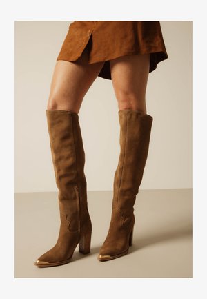 Bruine suède kniehoge laarzen met hoge houten hakken en gouden metalen tips, gedragen met een bruine rok op een neutrale achtergrond.