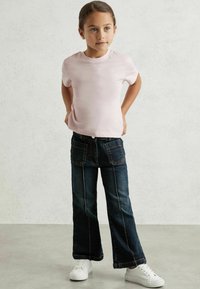 Lyserød t-shirt med korte ærmer parret med mørkeblå flared jeans, der har synlige sømme og to baglommer. Båret med hvide sneakers.