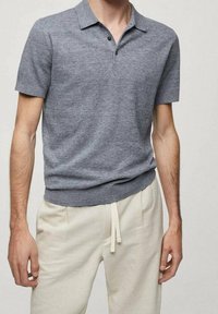Polo shirt gris à manches courtes avec une finition texturée, doté d'une patte de boutonnage et d'une taille ajustée. Porté avec un pantalon beige clair.