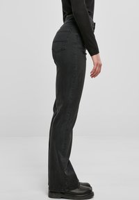 Vista lateral de una persona que lleva unos pantalones vaqueros negros de cintura alta, una camiseta negra de manga larga y botas negras, de pie frente a un fondo gris.