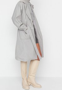 Trench-coat gris clair à coupe ample, manches longues et poches latérales. Associé à des bottes en caoutchouc beige montant jusqu'aux genoux.