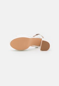 Sandal de tacón blanco con una suela sólida de color marrón claro, que presenta una correa en el tobillo y una superficie lisa. El tacón tiene una forma cuadrada para mayor estabilidad.