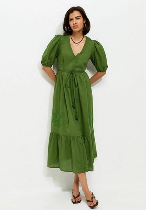 Kobieta stojąca w zielonej sukience midi z bufkami, dekoltem w serek, zapinanej na guziki z wiązaniem w talii i kwiatowym haftem, w połączeniu z czarnymi sandałami.