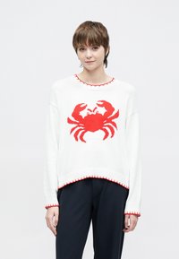 Λευκό πουλόβερ με κόκκινο σχέδιο καβουριού, ριμπ λαιμόκοψη και τελείωμα. Διαθέτει αντίθετη ραφή. Το μοντέλο φοράει μαύρο παντελόνι. Μαλακή υφή πλεκτού.