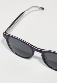 Ochelari de soare de culoare bleu marin cu lentile întunecate, având o dâră subțire roșie și albă în partea de sus și brațe metalice argintii. Rame din plastic neted.
