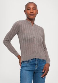 Sivý pletený kardigán s rebrovanou textúrou, so stojacím golierom a zapínaním na gombíky, skombinovaný s modrými džínsami.