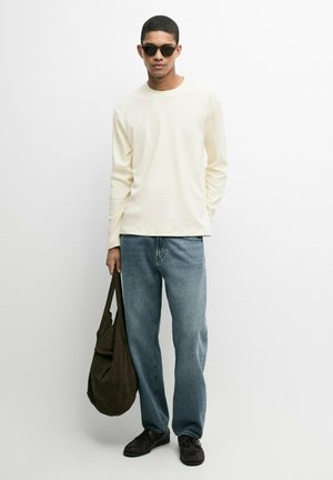 Uomo che indossa una camicia cremosa a maniche lunghe, jeans blu, scarpe scure e occhiali da sole, che tiene una grande borsa marrone su sfondo bianco.