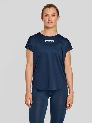 Vrouw met blond haar in een paardenstaart, gekleed in een donkerblauw sportshirt met korte mouwen en bijpassende leggings, staand tegen een effen grijze achtergrond.