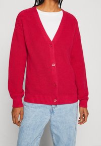 Kardigan v krvavě červené barvě s jemným žebrováním, dlouhými rukávy, ve tvaru V a pěti knoflíky. Měkká textura; má žebrované manžety a lem. Spárován s pastelově modrými džíny.