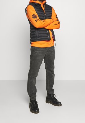Personne portant un sweat à capuche orange avec des motifs de roses noires sur les manches, un gilet matelassé noir, un jean gris foncé et des bottes noires à lacets, debout les bras croisés.