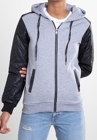 Sudadera con cremallera gris con mangas negras acolchadas, puños elásticos y capucha con cordón. Cuenta con dos bolsillos laterales y un acabado suave.