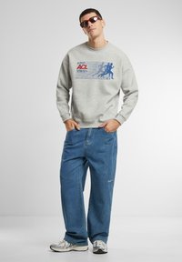 Hanorac gri deschis cu grafice albastre, asociat cu blugi lejeri albaștri. Modelul poartă ochelari de soare roșii și adidași argintii cu un design texturat.