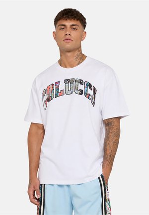Man with tattoos wearing a white t-shirt with colorful "COLLUCCI" text and light blue shorts with black vertical stripes and floral patterns.