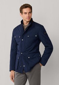 Marineblå quiltet jakke med høj krave, trykknapper, fire forlommer og tonale skulderdetaljer. Bæres over en grå skjorte.