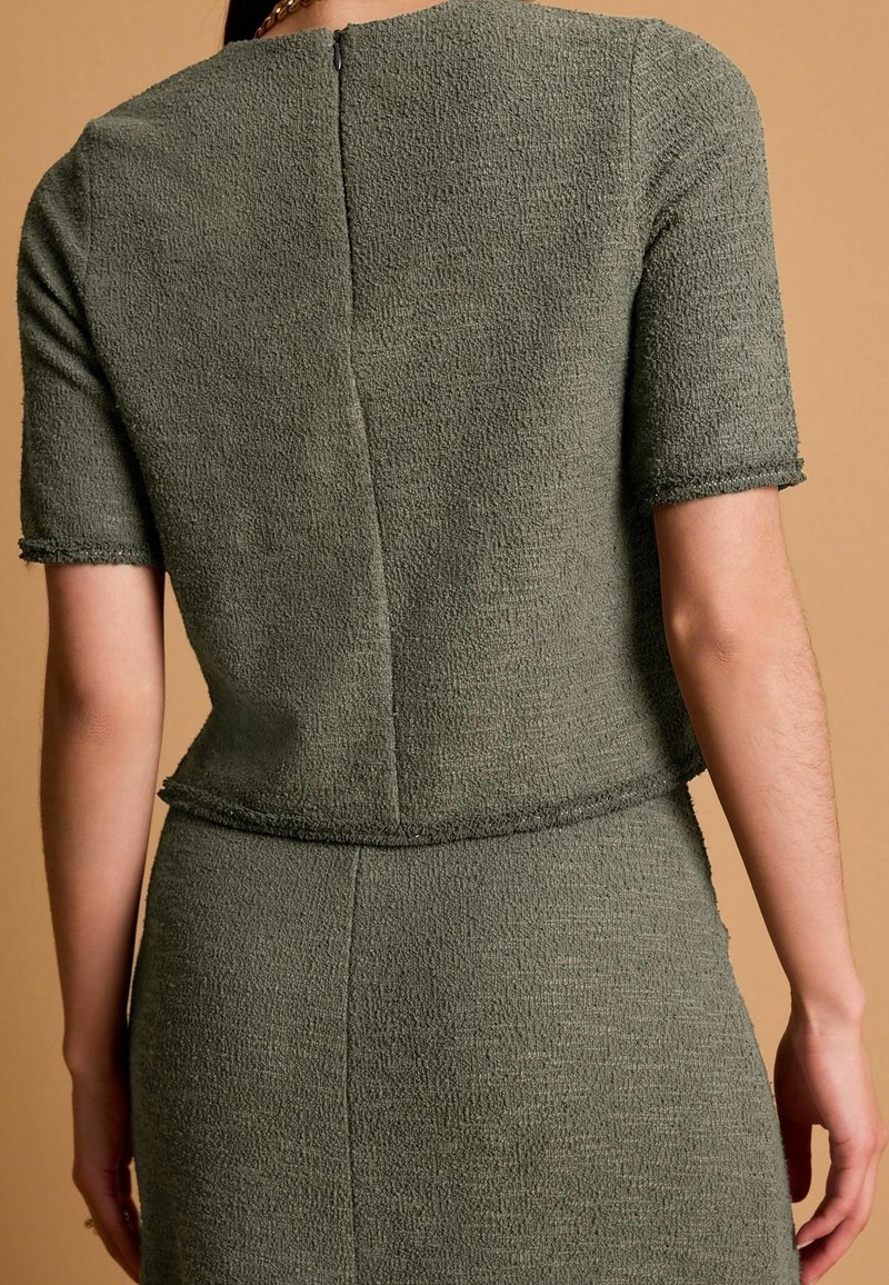 Femme portant un haut à manches courtes texturé vert olive et une jupe assortie, vue de dos sur un fond beige.