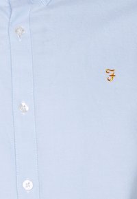 Helles blaues Baumwollhemd mit glatter Textur, ausgestattet mit einem kleinen in Gold gestickten Logo auf der Brust und klaren Knöpfen.