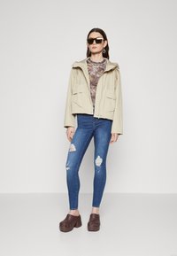 Beige kapucnis dzseki két elülső zsebbel, mintás felső, koptatott kék skinny farmernadrág és barna klumpák kivágásokkal és vastag sarokkal.