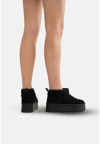 Botas de tobillo de ante negro con suela de plataforma, que presentan un diseño curvado, costuras laterales y una tira de ayuda en la parte trasera.