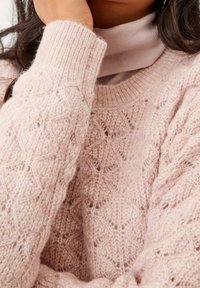Pull rose tricoté avec un motif texturé, des poignets côtelés et un col rond superposé à un haut à col roulé rose clair.