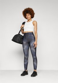 Leggings neri con motivi a onde blu, vita alta, abbinati a un top corto bianco. Una borsa a tracolla nera è appesa a una spalla.