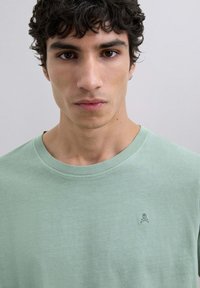 Camiseta de algodón verde menta con cuello redondo y un sutil logo de calavera y huesos cruzados en el pecho. Textura suave.