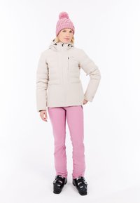Beige pufferjack met een ritssluiting aan de voorkant, schuine zakken en een afneembare capuchon, gecombineerd met roze skibroeken en zwarte skischoenen. Roze gebreide beanie.