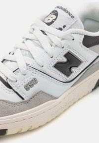 White leather sneaker with gray suede accents, mesh panels, black logo, and gray rubber sole. Features stitched detailing and lace closure.