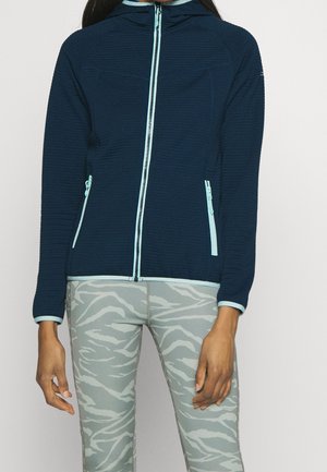 Kvinde iført en marineblå lynlåstrøje med lyseblå lynlåse og grå leggings med zebramønster.