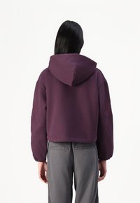 Felpa viola tagliata con cappuccio, dotata di maniche lunghe e dettagli testurizzati sulle spalle. Abbinata a pantaloni grigi.