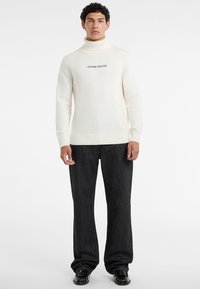 Maglione a collo alto crema con polsini e orlo a coste, con il logo "GUESS JEANS" in nero. Abbinato a jeans scuri a gamba dritta e scarpe lucide.