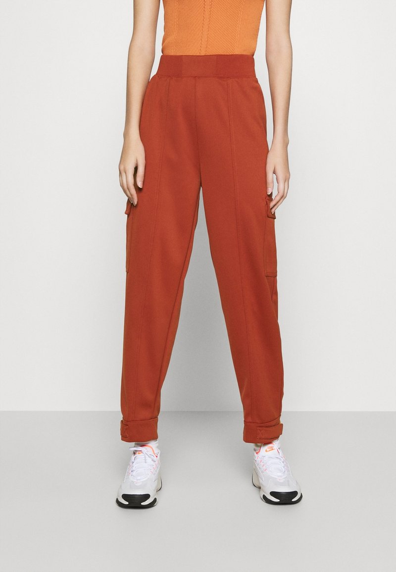 Cargo-byxor i rostorange, med hög midja, sidofickor och en tapered benavslutning med muddar. Matchas med vita och grå sneakers.