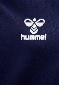 Marineblaue Stoff mit glatter Textur, der ein weißes Hummel-Logo mit einem stilisierten Bienen-Design und dem Markennamen darunter zeigt.